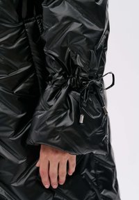 Zwarte pufferjack met een gestructureerde, glanzende afwerking. Beschikt over elastische manchetten met een trekkoord en metalen uiteinden, wat functionele details toevoegt.