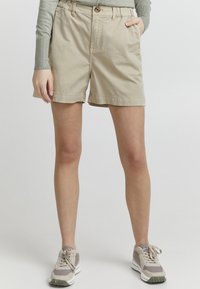 Beige bomullsshorts med knappeåpning, frontlomme og rullet kant. Kombinert med grå langermet topp og nøytrale sneakers.