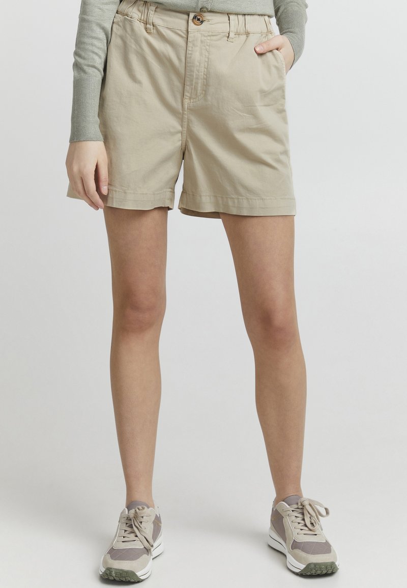 Beige bomullsshorts med knappeåpning, frontlomme og rullet kant. Kombinert med grå langermet topp og nøytrale sneakers.