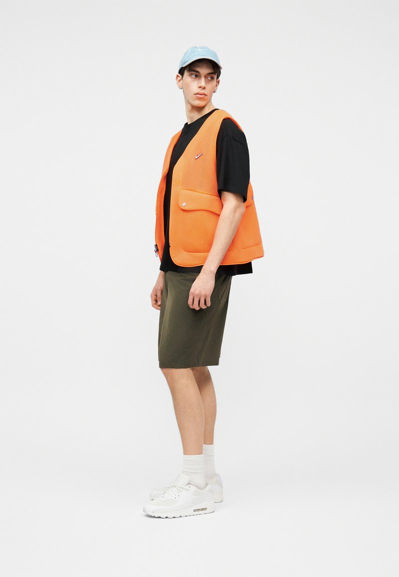 Junger Mann mit blauem Cap, schwarzem T-Shirt, orangefarbener Weste, olivgrünen Shorts, weißen Socken und weißen Turnschuhen, steht und schaut zur Seite.