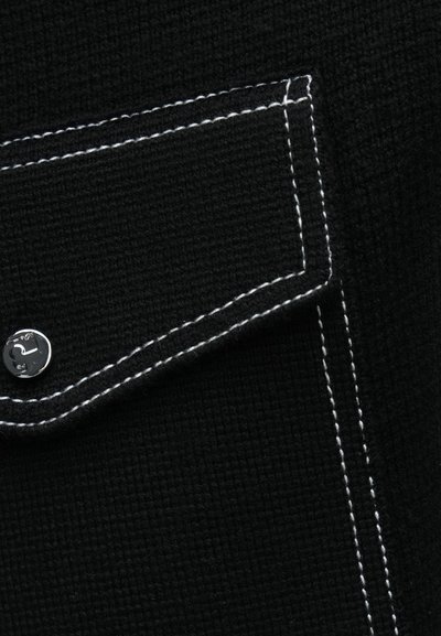 Mustast tekstuuriga kangas, millel on esiservas valgete kontrastsete õmblustega tasku ja metallist nööpkinnitusega detail.