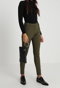 Olivgröna högmidjade byxor med avsmalnande ben, kombinerat med en svart långärmad topp. Håller en svart strukturerad klutch med ring. Svarta nitar skor.