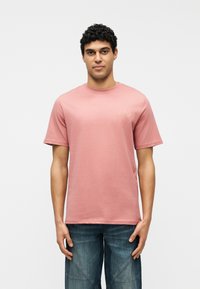 Άνδρας με σγουρά σκούρα μαλλιά, φορώντας ροζ κοντομάνικο μπλουζάκι και μπλε τζιν, στέκεται μπροστά σε απλό λευκό φόντο, κοιτώντας προς τα εμπρός.