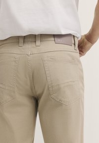 Beige strukturierte Hosen mit geradem Schnitt, ausgestattet mit zwei Gesäßtaschen und einem braunen Lederlogo-Patch am Bund.