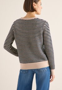 Gebreide sweater met een marineblauwe en crème stippenpatroon, geribbelde halslijn en onderste rand, en een relaxte pasvorm. Gdraagd met blauwe wijde jeans.