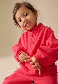 Pull polaire rose vif avec col à demi-fermeture éclair, poignets côtelés et pantalon assorti. Texture douce et coupe décontractée, adaptée aux enfants.