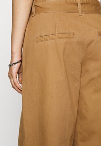 Bruna vida byxor med en slät textur, med en platt bakficka och en sydd midja. Ett silverarmband syns på handleden.