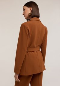 Blazer marrón a medida con cintura ceñida, hombros estructurados y mangas largas. Tejido suave con un diseño minimalista y sin patrones visibles.