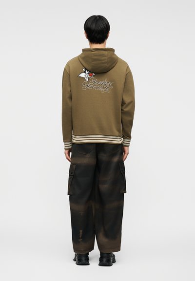 Άνδρας που φοράει φούτερ με κουκούλα σε ελιά χρώμα με χαρακτήρα κινουμένων σχεδίων και το κείμενο "Sufferin’ Succotash" στην πλάτη, μαύρο παντελόνι cargo και μαύρα παπούτσια, γυρισμένος πλάτη.