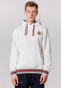 Hvid hoodie med en frontlomme, snørebånd i rød og marineblå, og et cirkulært logo-patch. Har stribede ribbede manchetter og kant.