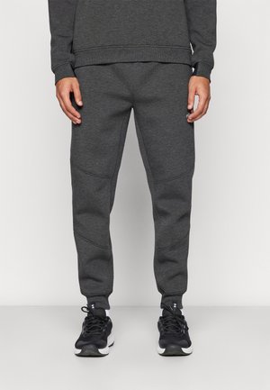 Mand iført mørkegrå joggebukser og matchende sweatshirt med sorte og hvide sportssko, stående mod en ensfarvet lys baggrund.