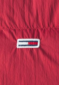 Tissu rouge avec une couture horizontale et un petit patch rectangulaire comportant des rayures horizontales blanches, bleu marine et rouges.