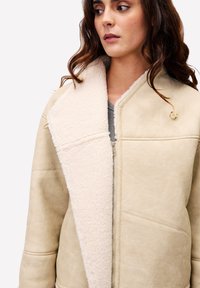 Vrouw in een beige shearling jack met grote kraag, rits aan de voorkant en houten knoopdetails, kijkend opzij.