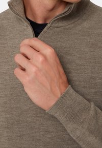 Ruka muže upravuje zip na béžovém pleteném svetru s žebrovanými manžetami přes černé tričko.