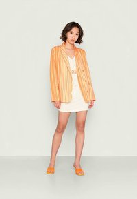 Blazer listrado em laranja e branco com lapela em bico e botões duplos, usado sobre um vestido creme. Modelo calça sandálias slides laranja.