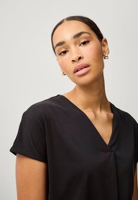 Sort kortærmet top med V-udskæring, lavet af glat stof og med en afslappet pasform. Modellen har guld ringe i ørerne og naturlig makeup.