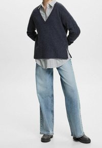 Dunkelblauer V-Ausschnitt-Pullover mit gerippter Textur, über einem weiß-blau gestreiften Hemd getragen, kombiniert mit lässigen hellblauen Jeans und schwarzen Schuhen.