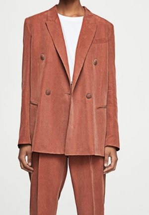 Veste croisée en velours côtelé couleur rouille. Caractéristiques : grands boutons, revers à cran et poches latérales. Associée à un pantalon assorti.