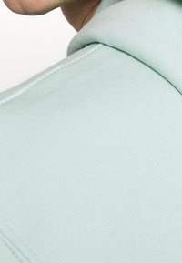 Sudadera verde menta con una textura suave, que presenta una costura visible a lo largo del borde y un corte relajado. Primer plano del área del cuello en la parte posterior.