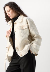 Helles, strukturiertes Jacket mit Fransenakzenten, goldenen Knöpfen und zwei Brusttaschen, über einem schwarzen Oberteil getragen und mit schwarzen Hosen kombiniert.