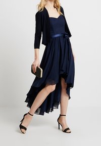 Žena v tmavě modrých šatech s vysokým a nízkým lemem, černém krátkém kardiganu, černých sandálech na kotníku, držící malou kabelku se zlatými akcenty.