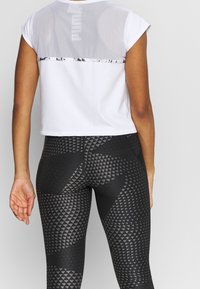 Top blanco corto con espalda de malla y herrajes plateados, combinado con leggings negros que presentan un patrón geométrico de diamantes.