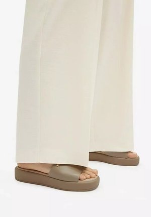 Füße mit beigen Plateau-Sandalen kombiniert mit weitem, offwhite Hosen vor weißem Hintergrund.