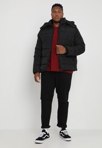 Sort pufferjakke med lynlås foran og hætte, med quiltet polstring. Bæret over en rød skjorte og parret med sorte bukser og sorte sneakers.