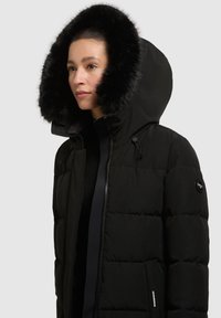 Schwarze Pufferjacke mit abnehmbarem schwarzen Kunstfellkragen. Verfügt über einen Reißverschluss, gestepptes Design und Logostick auf dem linken Arm.