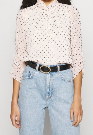 Kvinde iført lyserød bluse med små sorte print, stukket ned i lyseblå højtsiddende jeans med sort bælte og guldlås.