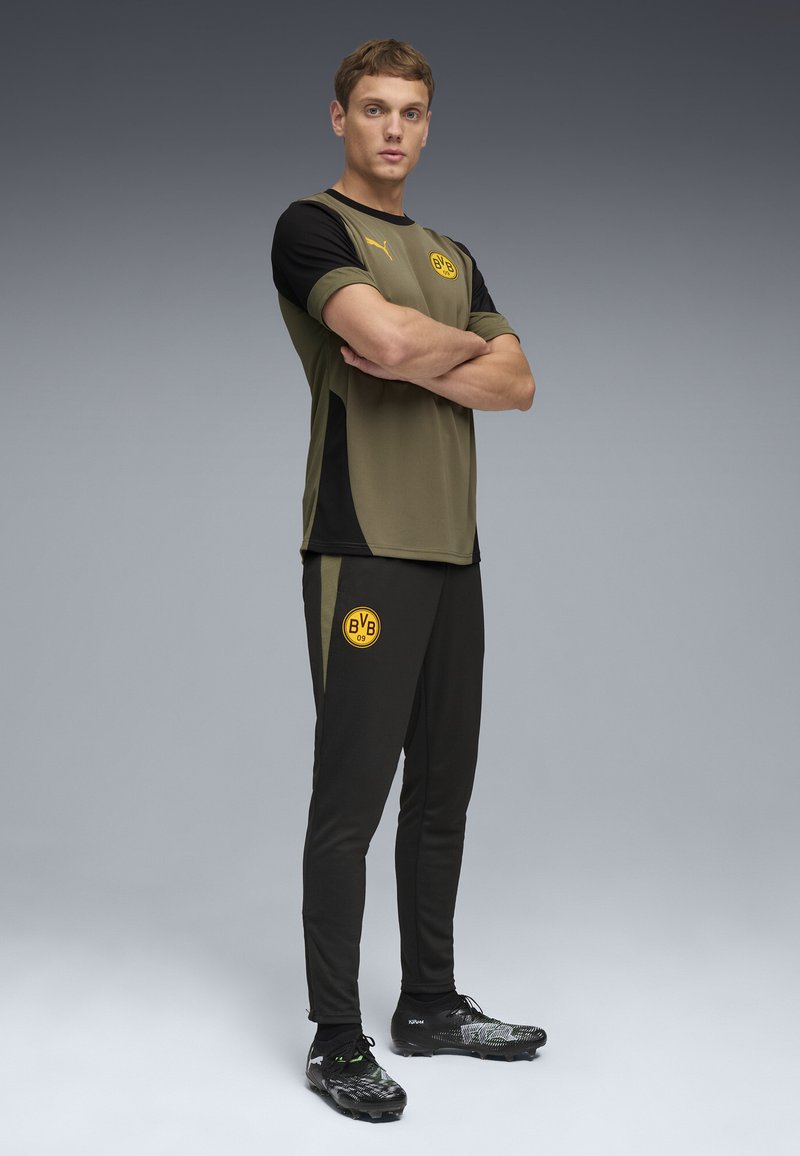 Junger männlicher Athlet steht mit verschränkten Armen, trägt ein olivgrünes und schwarzes Borussia Dortmund Trainingsoutfit und schwarze Stollenschuhe vor grauem Hintergrund.