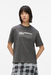 Camiseta de algodón gris con mangas cortas, cuello redondo y texto en blanco "KARL LAGERFELD JEANS" impreso en el frente. La lleva puesta con una falda a cuadros.