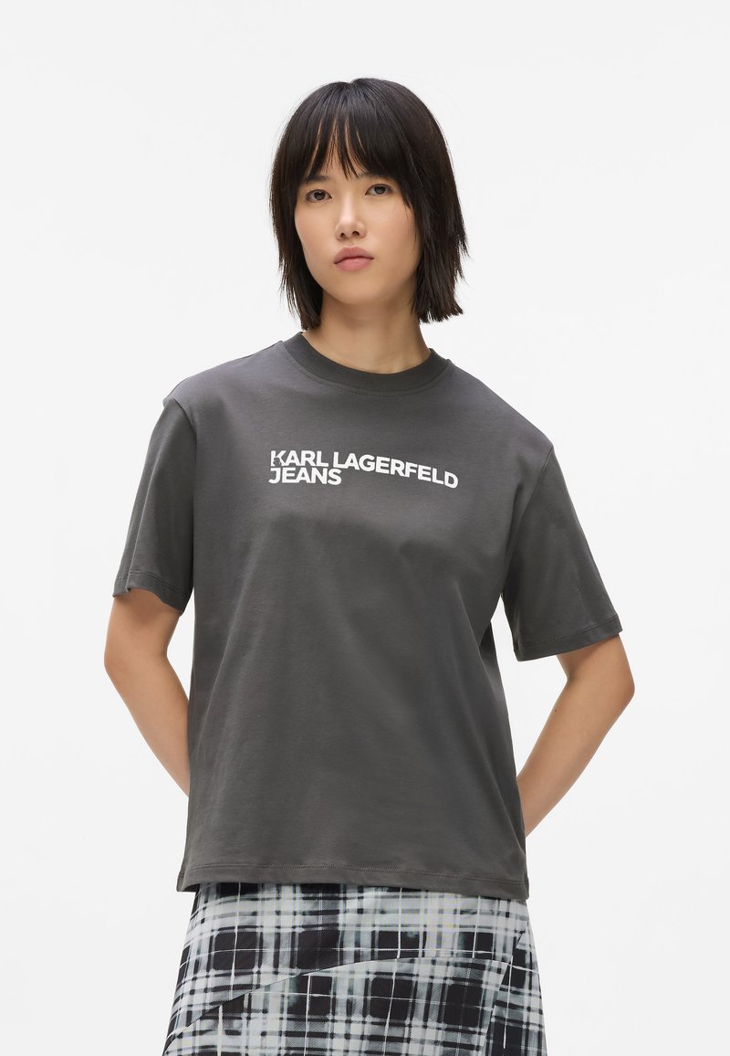 Camiseta de algodón gris con mangas cortas, cuello redondo y texto en blanco "KARL LAGERFELD JEANS" impreso en el frente. La lleva puesta con una falda a cuadros.