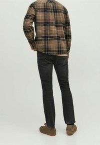 Man som bär en brun och svart rutigt skjorta, svarta jeans och bruna mockaskor, står med ryggen mot kameran.