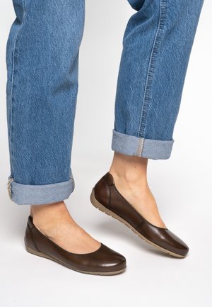 Brown leather flat shoes with a rounded toe, textured finish, and a flexible beige sole, paired with rolled-up blue denim pants.