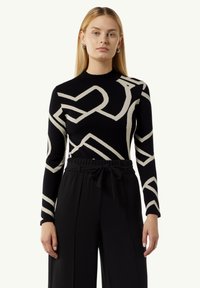 Schwarzer Strickpullover mit cremefarbenem abstraktem Muster, runder Ausschnitt und enganliegenden langen Ärmeln. Kombiniert mit hochtaillierten schwarzen Hosen mit Bindegürtel.