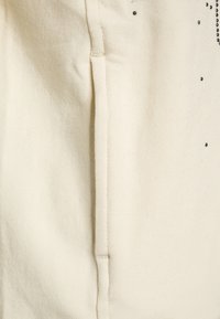 Tessuto crema con una cucitura verticale e piccoli punti decorativi neri sparsi vicino all'angolo in alto a destra.