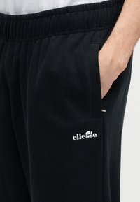 Κοντινή λήψη μαύρων φόρμας με ελαστική μέση, χέρι στην πλευρική τσέπη και λευκό λογότυπο ellesse στον μηρό.