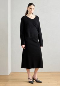 Čierny pletený sveter s voľným strihom a výstrihom do V, v kombinácii s čiernou rebrovanou midi sukňou. Nosené s šedými slingback podpätkami na drevenej podlahe.