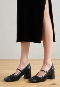 Black heeled shoes with a woven upper and smooth leather accents, featuring a buckle strap and a square block heel.