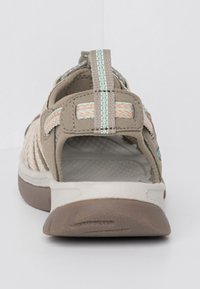 Light brown sandal with woven straps in peach and teal, cushioned footbed, and a thick white sole. Rear strap for secure fit.