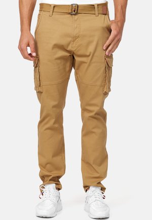 Man draagt beige cargobroek met zijzakken, een beige riem en witte sneakers, staand voor een effen achtergrond.