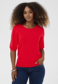 Rød kortærmet sweater med rund halsudskæring, glat tekstur og en tætsiddende silhuet, parret med mørkeblå jeans. Ingen bemærkelsesværdige mønstre.