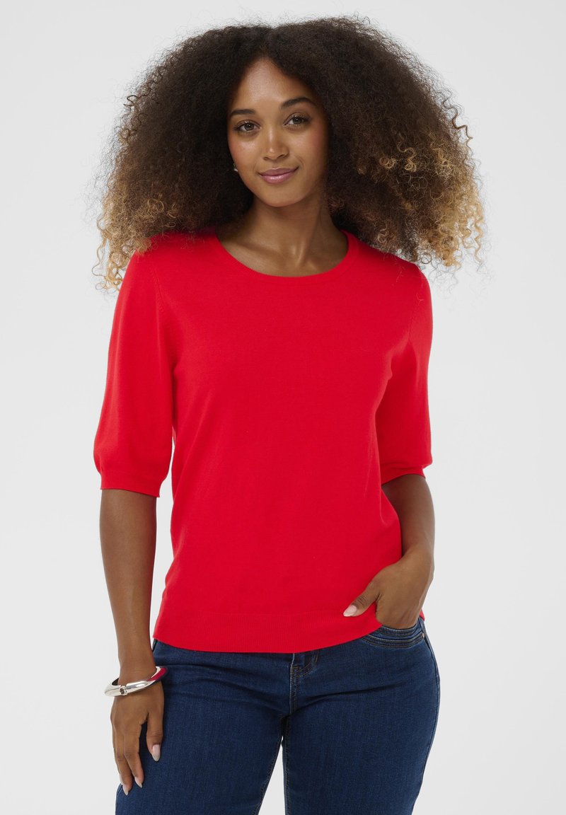 Rød kortærmet sweater med rund halsudskæring, glat tekstur og en tætsiddende silhuet, parret med mørkeblå jeans. Ingen bemærkelsesværdige mønstre.