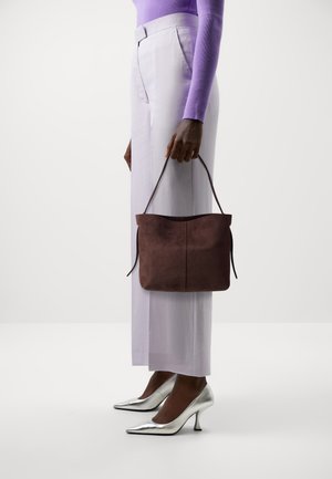Vrouw in lila top en witte broek met een bruine suède handtas, zilveren hoge hakken, staand tegen een effen achtergrond.
