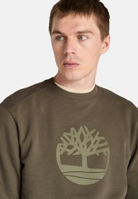 Sudadera de color verde oliva con un gran logo de árbol de color verde claro en el frente. Presenta un cuello redondo y una tela texturada. Diseño casual y sin adornos.