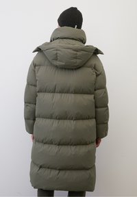 Person wearing long olive-green padded winter coat with hood and black knit beanie, facing away against plain light background.