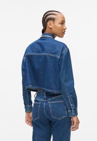 Giacca di jeans cropped di colore blu scuro, con colletto alla coreana, dettagli cuciti e maniche lunghe con polsini abbottonati.