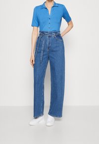 Blue short-sleeve polo shirt with a collar and button placket, paired with high-waisted, wide-leg blue denim jeans and white sneakers.
