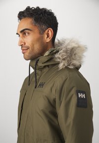 Maslinasto zeleni parka kaput s krznom obloženim kapuljačom, džepovima na zips i logotipom na rukavu. Vodootporan materijal s glatkom teksturom.